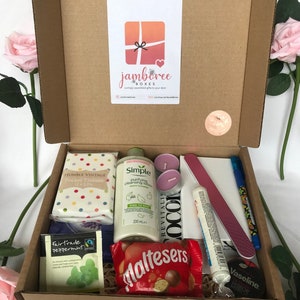 May include: A brown cardboard box filled with a variety of gifts, including a bottle of Simple Purifying Cleansing Lotion, a pack of Humble Vintage Pocket Tissues, a box of Maltesers chocolates, a pack of Fairtrade Peppermint tea, two pink candles, a nail file, a tube of Vaseline lip balm, and a blue pen.