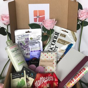 May include: A brown cardboard box filled with various items, including a pink face mask, a bottle of face wash, a tube of Vaseline, a pack of tissues, a pack of Maltesers, two pink candles, two tea bags, a purple nail file, a blue pen, and a pink and white foot scrub. The box also contains two pink roses and a small card with a red bow.