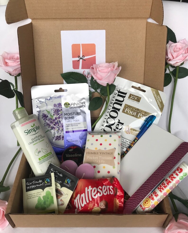 May include: A brown cardboard box filled with various items, including a pink face mask, a bottle of face wash, a tube of Vaseline, a pack of tissues, a pack of Maltesers, two pink candles, two tea bags, a purple nail file, a blue pen, and a pink and white foot scrub. The box also contains two pink roses and a small card with a red bow.