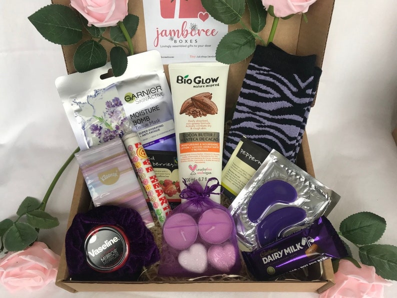 May include: A brown cardboard box filled with a variety of self-care items, including a face mask, body lotion, socks, eye masks, candles, and chocolate. The box is decorated with a pink rose and the text "jamboree boxes".