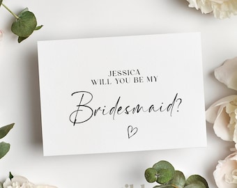 May include: A white card with the text "JESSICA WILL YOU BE MY Bridesmaid?" in black script, with a small heart below. The card is surrounded by white peonies and green eucalyptus leaves, with the Wild Bloom logo at the bottom.