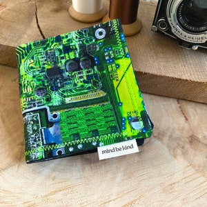 May include: A folded wallet featuring a green circuit board design. The wallet has a white label with the text "mind be kind". White thread stitches the edges. The background includes a wooden surface, thread spools, and a vintage camera.