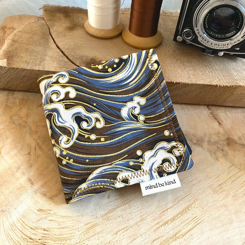 May include: Square fabric item with a wave pattern in shades of brown, blue, white, and gold. The fabric is folded and has a small white tag that reads "mind be kind". The item is likely a handkerchief or similar accessory.
