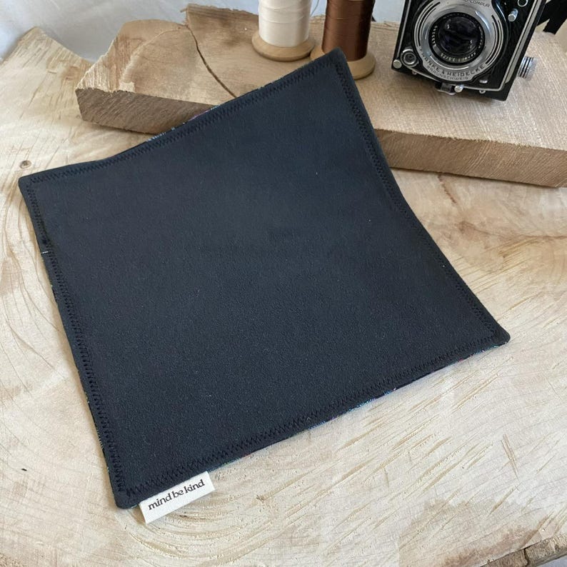 May include: A square, black fabric coaster with a stitched edge. The coaster has a small white tag with the text "mind be kind" in a simple font. The coaster is displayed on a wooden surface, with a vintage camera and spools of thread in the background.