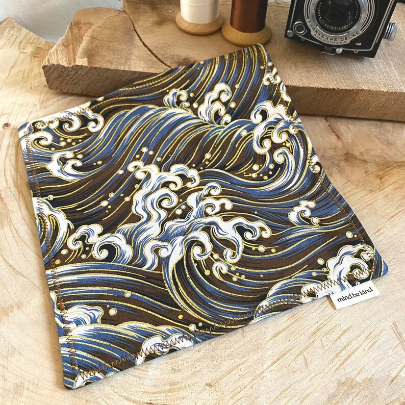 May include: Square fabric item with a repeating wave pattern in shades of brown, blue, and gold. The design features stylized waves and foam. The item has a white tag that reads "mind be kind".