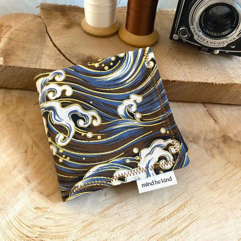 May include: Square fabric item with a wave pattern in shades of blue, brown, and gold. The fabric has a white label that reads "mind be kind". The item is likely a handkerchief or small cloth.