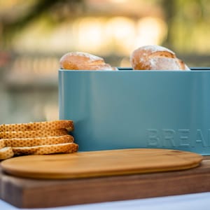 Puede incluir: Una panera turquesa con la palabra "BREAD" en relieve en la parte delantera, que contiene dos panes. Pan en rodajas y una tabla de cortar de madera se encuentran sobre una tabla de madera más grande. La escena está ambientada al aire libre.
