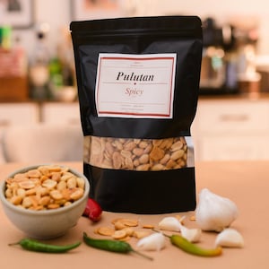May include: A black stand-up pouch labeled Pulutan Spicy containing roasted peanuts, with a small bowl of peanuts, fresh garlic cloves, and green chili peppers arranged in front of the packaging on a flat surface.