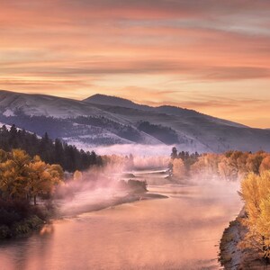 May include: A scenic landscape photograph of a river winding through a valley at sunrise. The sky is a gradient of orange and pink, with mountains in the background and trees with yellow foliage along the riverbanks. Mist hangs over the water.