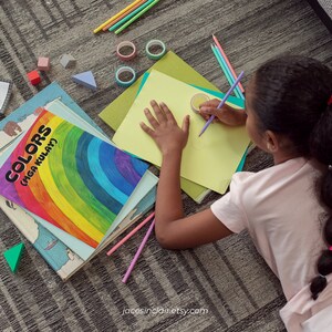 Op de afbeelding: Een jong persoon zit op de vloer en tekent op een vel geel papier met een paarse potlood. Ze zijn omringd door kleurpotloden, kleurpotloden en een boek met de titel "Colors (Mga Kulay)".