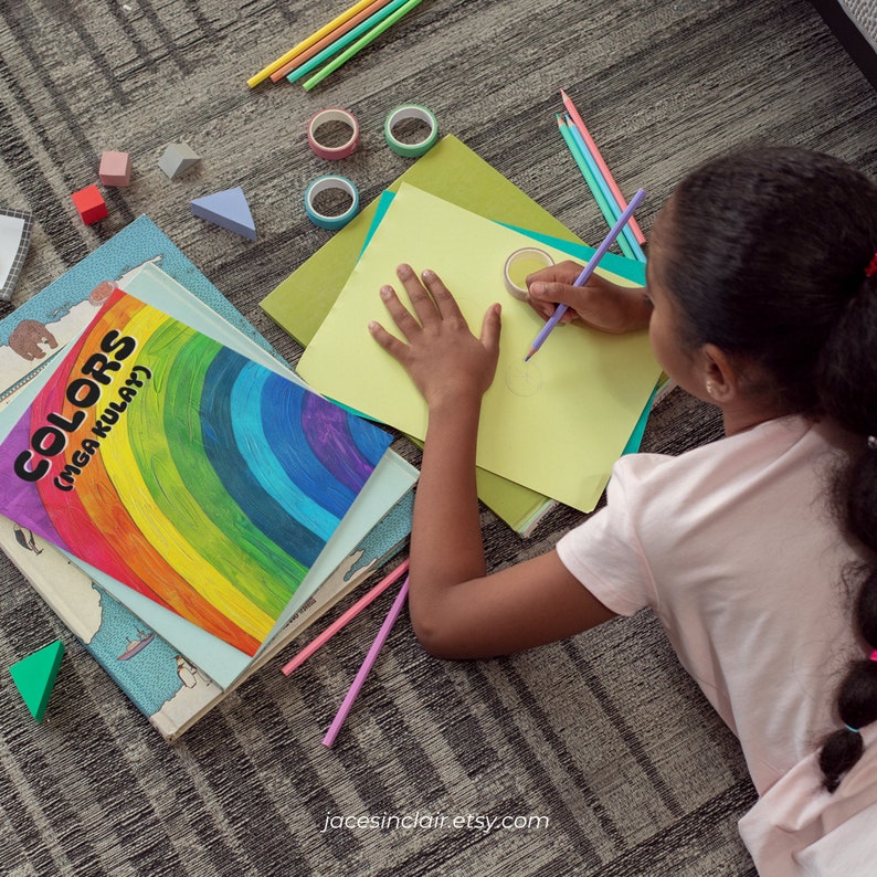 Op de afbeelding: Een jong persoon zit op de vloer en tekent op een vel geel papier met een paarse potlood. Ze zijn omringd door kleurpotloden, kleurpotloden en een boek met de titel "Colors (Mga Kulay)".