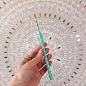 May include: A fine-tipped, mint green artist's brush with a silver ferrule and a red bristle tip. The brush is held against a backdrop of a white, circular, ornate decorative piece with mirrored accents.