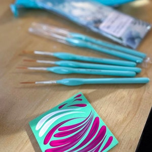 Op de afbeelding: Een set van zes turquoise verfkwasten met witte haren, gelegd op een houten oppervlak. Een klein vierkant stuk papier met een groene achtergrond en roze en witte swirls is op de voorgrond. Het papier heeft de tekst "@loveanddots" erop.