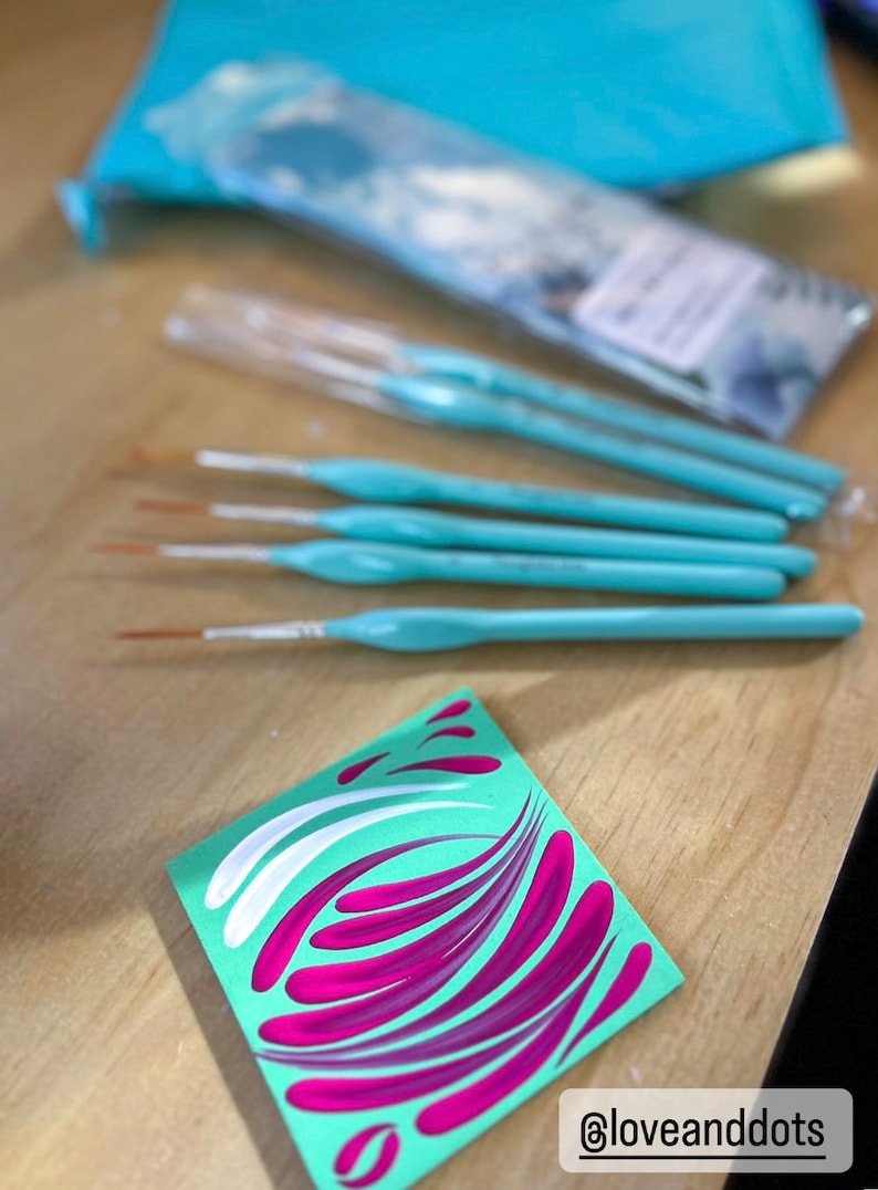 Op de afbeelding: Een set van zes turquoise verfkwasten met witte haren, gelegd op een houten oppervlak. Een klein vierkant stuk papier met een groene achtergrond en roze en witte swirls is op de voorgrond. Het papier heeft de tekst "@loveanddots" erop.