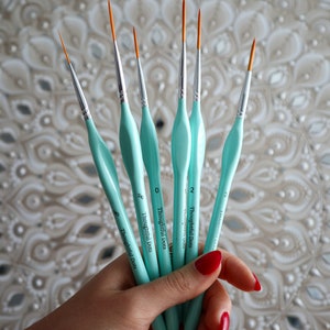 Op de afbeelding: Een set van zes turquoise verfkwasten met oranje punten. De kwasten zijn gelabeld "Thoughtful Dots" en hebben de maten 0, 2, 3, 4, 5 en 6.