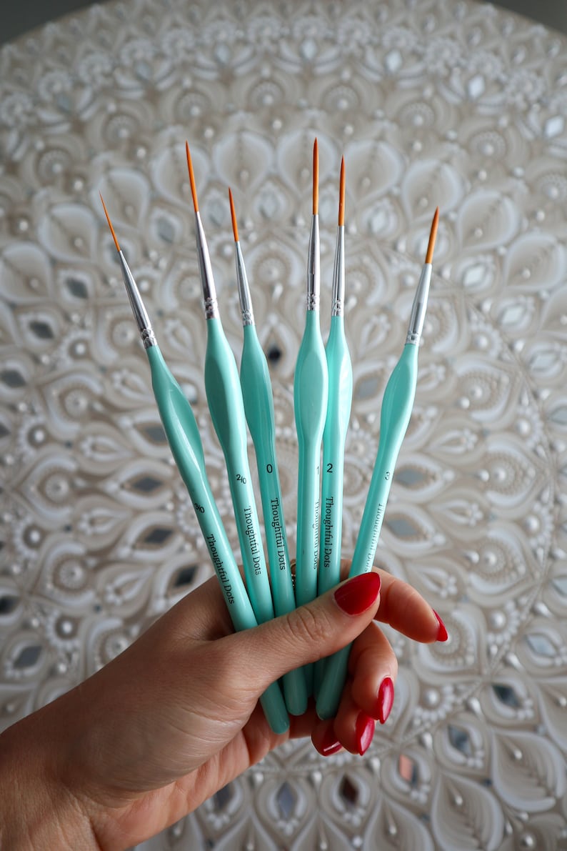 Op de afbeelding: Een set van zes turquoise verfkwasten met oranje punten. De kwasten zijn gelabeld "Thoughtful Dots" en hebben de maten 0, 2, 3, 4, 5 en 6.