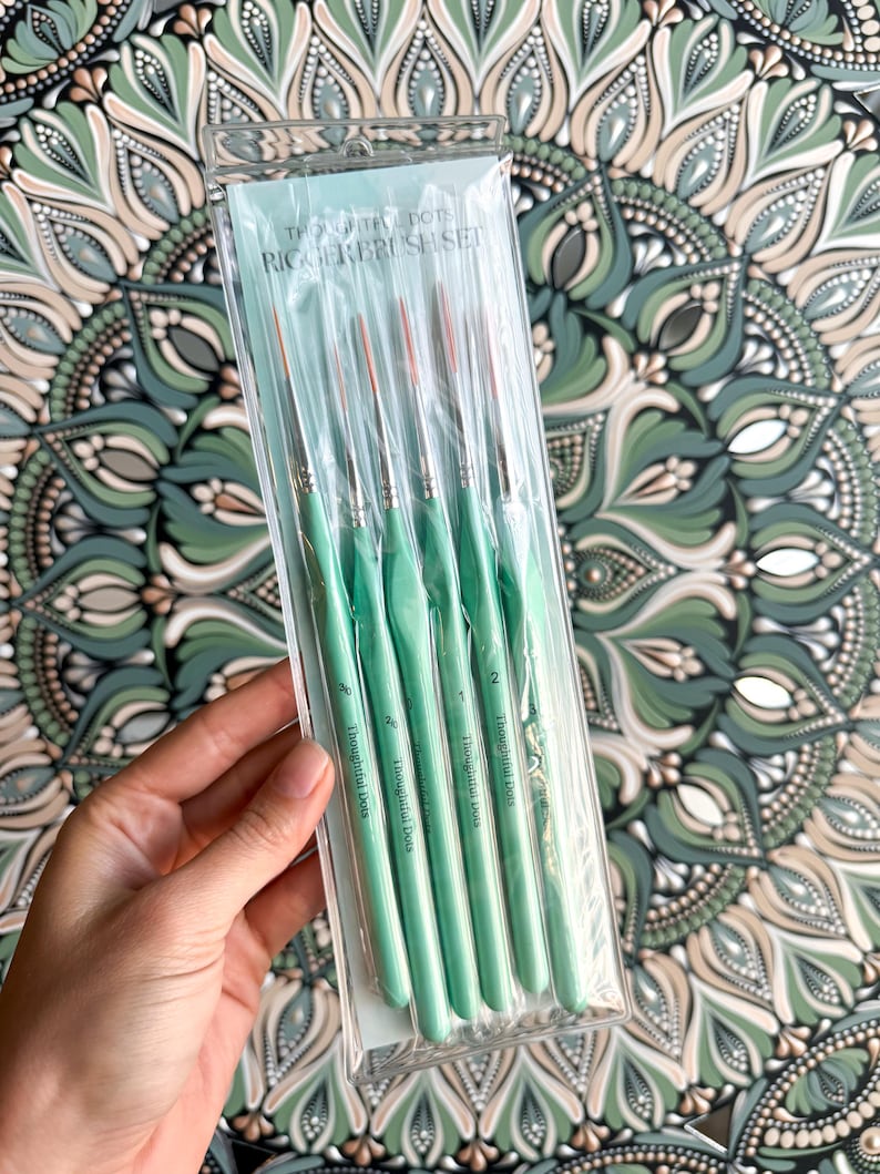 Op de afbeelding: Een set van zes groene verfkwasten met de tekst "Thoughtful Dots" en "Rigger Brush Set" op de verpakking. De kwasten zijn verpakt in een doorzichtige plastic hoes.