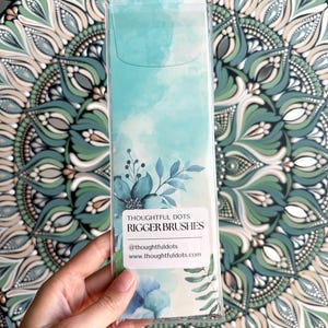 Op de afbeelding: Een transparante plastic verpakking met een set blauwe en groene aquarel-riggerkwasten. De verpakking heeft een wit label met de tekst "Thoughtful Dots Rigger Brushes" en de social media handle "@thoughtfuldots" en website "www.thoughtfuldots.com".