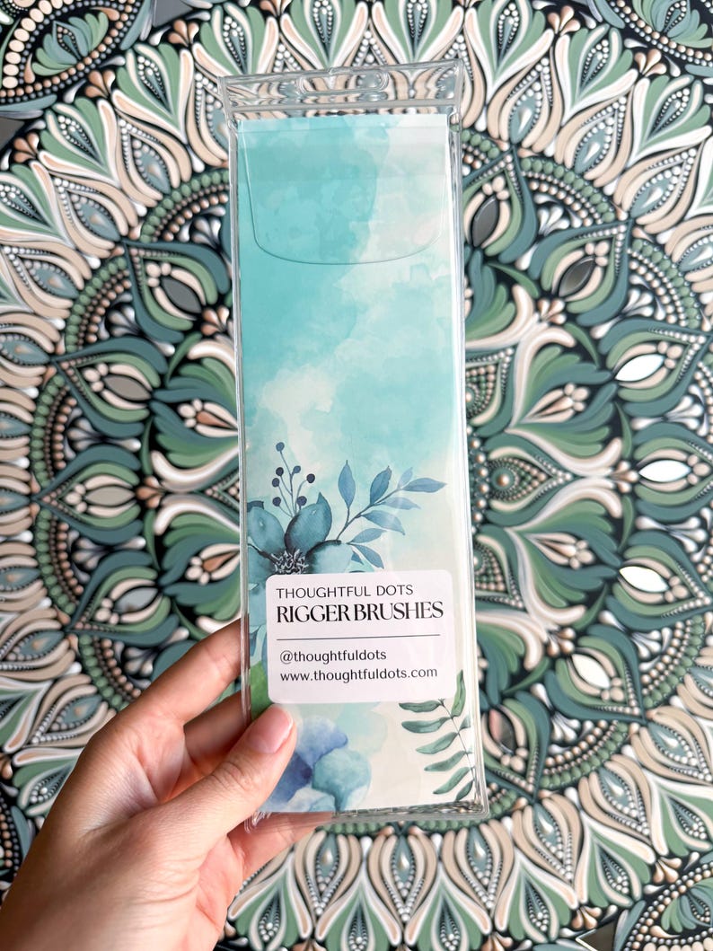 Op de afbeelding: Een transparante plastic verpakking met een set blauwe en groene aquarel-riggerkwasten. De verpakking heeft een wit label met de tekst "Thoughtful Dots Rigger Brushes" en de social media handle "@thoughtfuldots" en website "www.thoughtfuldots.com".
