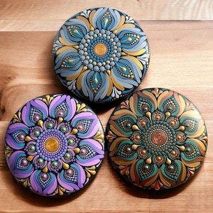 May include: Three round painted stones with mandala designs. The first stone has a blue and gold mandala with a white background. The second stone has a purple and gold mandala with a black background. The third stone has a green and gold mandala with a black background.