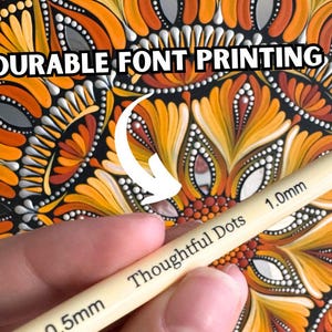 May include: A close-up of a hand holding a set of two dotting tools for creating dot art. The tools are labeled "Thoughtful Dots" and have a 0.5mm and 1.0mm tip. The background is a colorful mandala pattern with orange, yellow, and red colors.