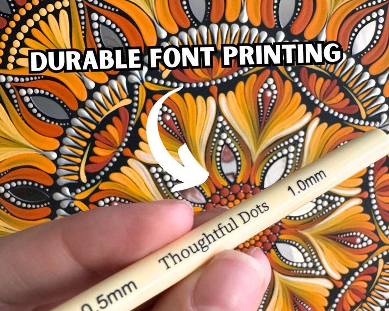 May include: A close-up of a hand holding a set of two dotting tools for creating dot art. The tools are labeled "Thoughtful Dots" and have a 0.5mm and 1.0mm tip. The background is a colorful mandala pattern with orange, yellow, and red colors.