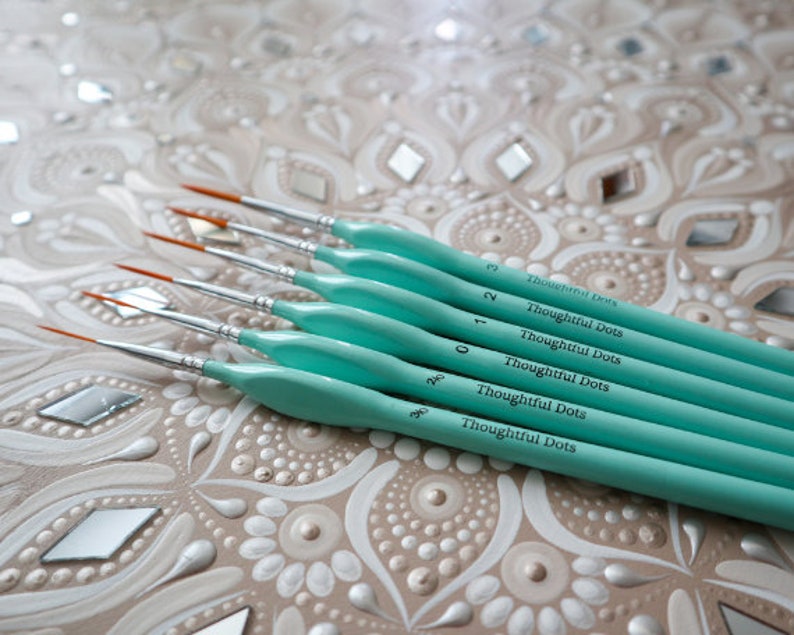 Op de afbeelding: Een set van zeven turquoise verfkwasten met zwarte letters die "Thoughtful Dots" en de nummers 0, 1, 2, 3, 2/0, 3/0 lezen. De kwasten zijn in een waaier op een wit en bruin gepatroneerd oppervlak gerangschikt.