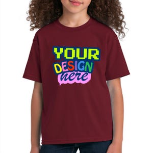 Può includere: Una t-shirt a maniche corte color bordeaux con una stampa grafica multicolore sul petto. Il testo recita "YOUR" in verde lime, "DESIGN" in giallo e "here" in un carattere corsivo rosa chiaro, il tutto su uno sfondo blu scuro.