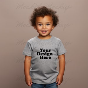 K&ouml;nnte beinhalten: Ein kleines Kind tr&auml;gt ein hellgraues T-Shirt mit dem Aufdruck "Your Design Here" auf der Vorderseite. Das Kind schaut direkt in die Kamera und l&auml;chelt.