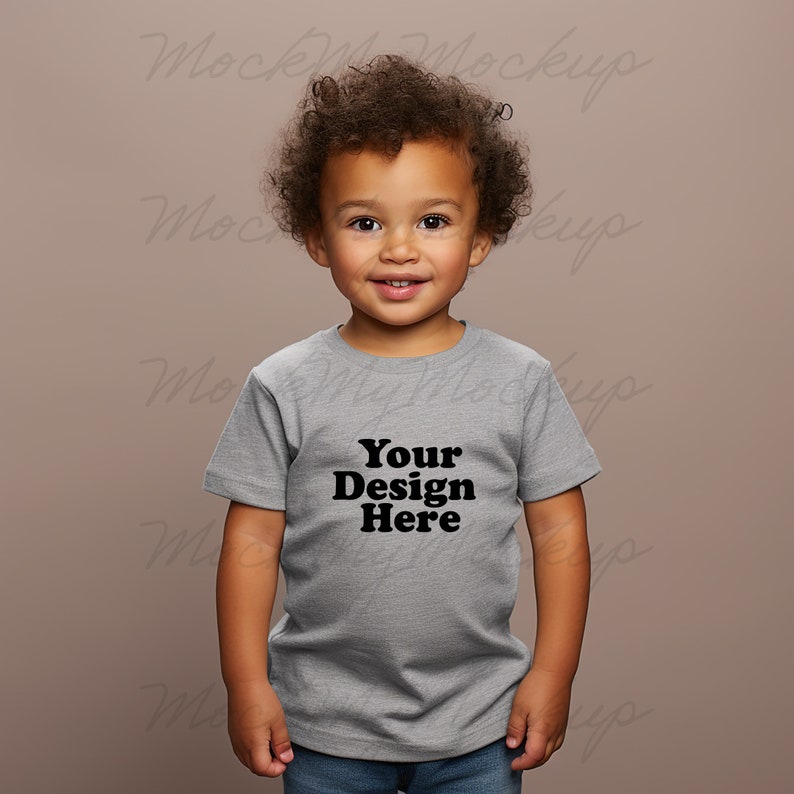 K&ouml;nnte beinhalten: Ein kleines Kind tr&auml;gt ein hellgraues T-Shirt mit dem Aufdruck "Your Design Here" auf der Vorderseite. Das Kind schaut direkt in die Kamera und l&auml;chelt.