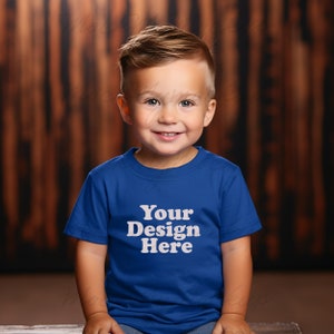 K&ouml;nnte beinhalten: Ein kleines Kind tr&auml;gt ein k&ouml;nigsblaues T-Shirt mit dem Text "Your Design Here" in Wei&szlig;. Das Kind sitzt vor einem Holz Hintergrund.