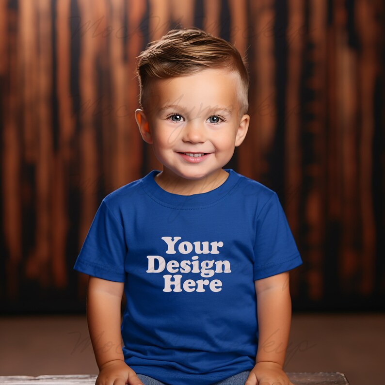K&ouml;nnte beinhalten: Ein kleines Kind tr&auml;gt ein k&ouml;nigsblaues T-Shirt mit dem Text "Your Design Here" in Wei&szlig;. Das Kind sitzt vor einem Holz Hintergrund.
