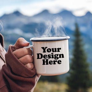 Könnte beinhalten: Eine weiße Emailletasse mit schwarzem Rand und Henkel. Die Tasse hat den Text "Your Design Here" in Schwarz auf der Vorderseite aufgedruckt. Die Tasse ist mit einem heißen Getränk gefüllt und Dampf steigt aus der Oberseite auf.