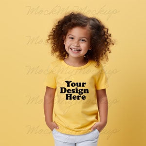 K&ouml;nnte beinhalten: Ein junges Kind tr&auml;gt ein gelbes T-Shirt mit dem Text "Your Design Here" in Schwarz. Das Kind l&auml;chelt und hat lockiges braunes Haar.