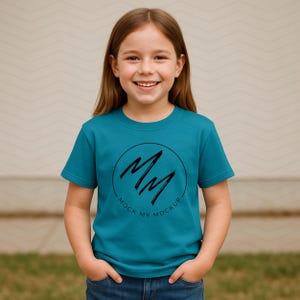 May include: A teal t-shirt with a black circular logo featuring the letters "MM" and the text "MOCK MY MOCKUP". The shirt is worn by a young person with long brown hair and blue jeans.