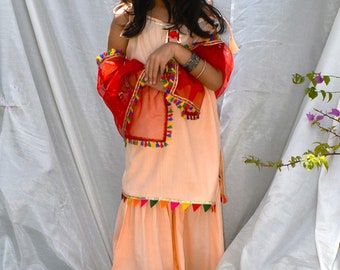 Könnte beinhalten: Ein junges Mädchen trägt ein pfirsichfarbenes Salwar Kameez mit bunten Stickereien und einen roten Dupatta. Sie trägt ein Juwelen-Stirnband und ein silbernes Armband.