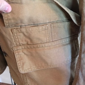 May include: A brown canvas jacket with a large flap pocket on the front. The pocket has two smaller pockets inside.