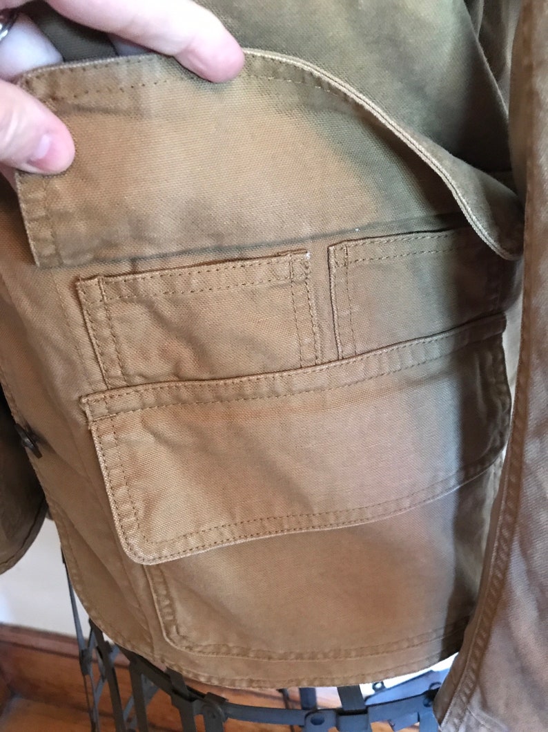 May include: A brown canvas jacket with a large flap pocket on the front. The pocket has two smaller pockets inside.