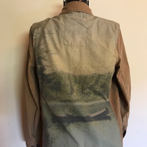 May include: A brown and green utility jacket with a faded image of a landscape on the back. The jacket has a button-up front and long sleeves.