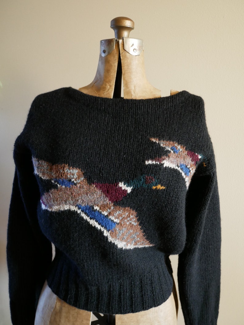 Pode incluir: Camisola preta de malha com gola redonda e desenho de pato. Os patos s&atilde;o retratados em tons de castanho, azul, branco e bord&ocirc;. A camisola &eacute; exibida em um manequim vintage.