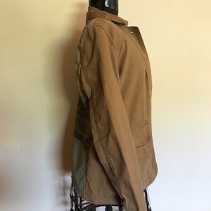 May include: A brown utility jacket with a camouflage pattern on the back. The jacket has a button closure and two pockets.