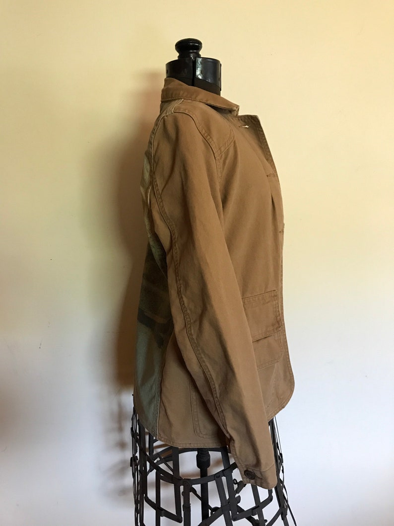 May include: A brown utility jacket with a camouflage pattern on the back. The jacket has a button closure and two pockets.