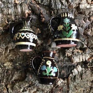 May include: Three small, decorative ceramic pitchers with handles. The pitchers are dark brown with hand-painted floral designs in blue, yellow, and white. One pitcher has a white and brown geometric pattern. The pitchers are displayed on a textured, brown surface.