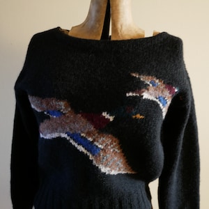 Pode incluir: Camisola preta de malha com decote barco e mangas compridas. A camisola apresenta um design abstrato colorido no peito, com detalhes em castanho, azul, branco e bord&ocirc;. A camisola &eacute; exibida em um manequim.