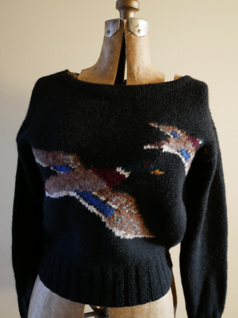 Pode incluir: Camisola preta de malha com decote barco e mangas compridas. A camisola apresenta um design abstrato colorido no peito, com detalhes em castanho, azul, branco e bord&ocirc;. A camisola &eacute; exibida em um manequim.