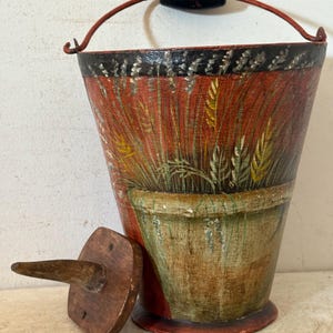 May include: A tapered bucket with a red base and a black rim, featuring a painted design of wheat stalks and a green pot. The bucket has a red metal handle with a black wooden grip and is accompanied by a separate, small, square wooden lid with a handle.