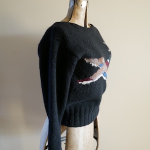 Pode incluir: Camisola preta de malha com gola redonda e mangas compridas. A camisola apresenta um design geom&eacute;trico colorido na frente, com tons de castanho, branco, azul e bord&ocirc;. A camisola &eacute; exibida em um manequim vintage.