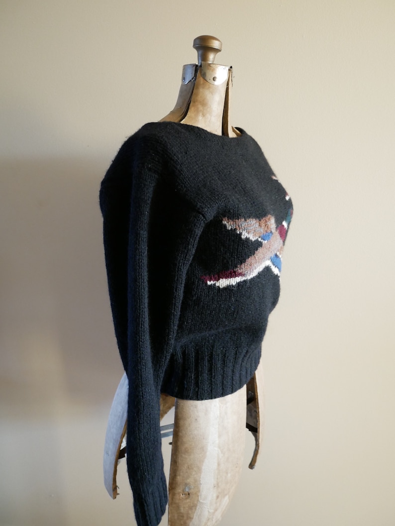 Pode incluir: Camisola preta de malha com gola redonda e mangas compridas. A camisola apresenta um design geom&eacute;trico colorido na frente, com tons de castanho, branco, azul e bord&ocirc;. A camisola &eacute; exibida em um manequim vintage.