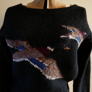 Pode incluir: Um su&eacute;ter preto de malha com decote barco e mangas compridas. O su&eacute;ter apresenta um design abstrato colorido na frente, com detalhes em marrom, azul, branco e bord&ocirc;. O su&eacute;ter &eacute; exibido em um manequim.