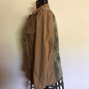 May include: A brown utility jacket with a camouflage pattern on the back. The jacket has a button closure and two pockets on the front.